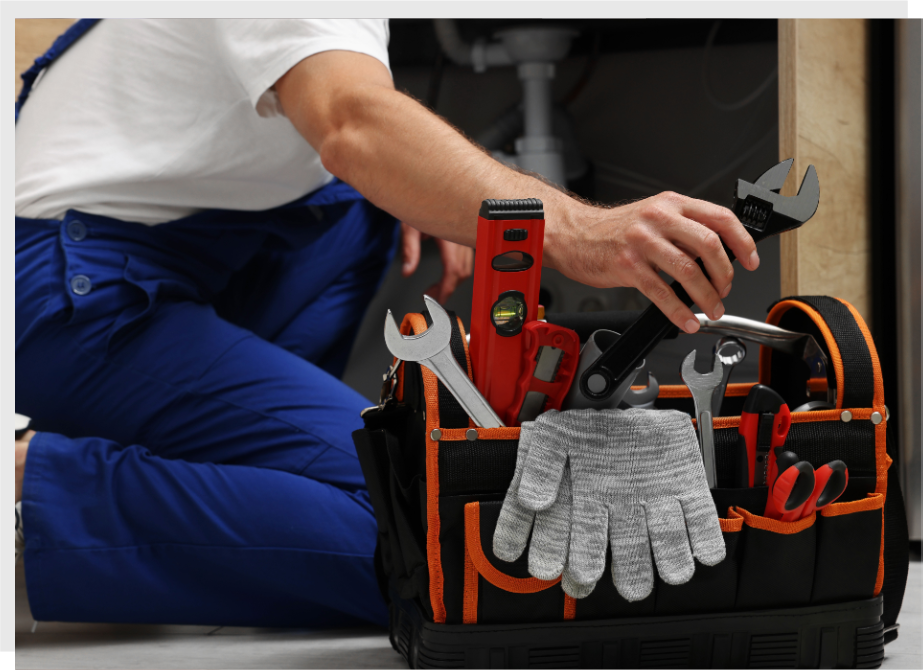 Person in blue overalls kneeling by a toolbox, selecting a wrench. The toolbox contains various tools like a level, gloves, pliers, and screwdrivers.