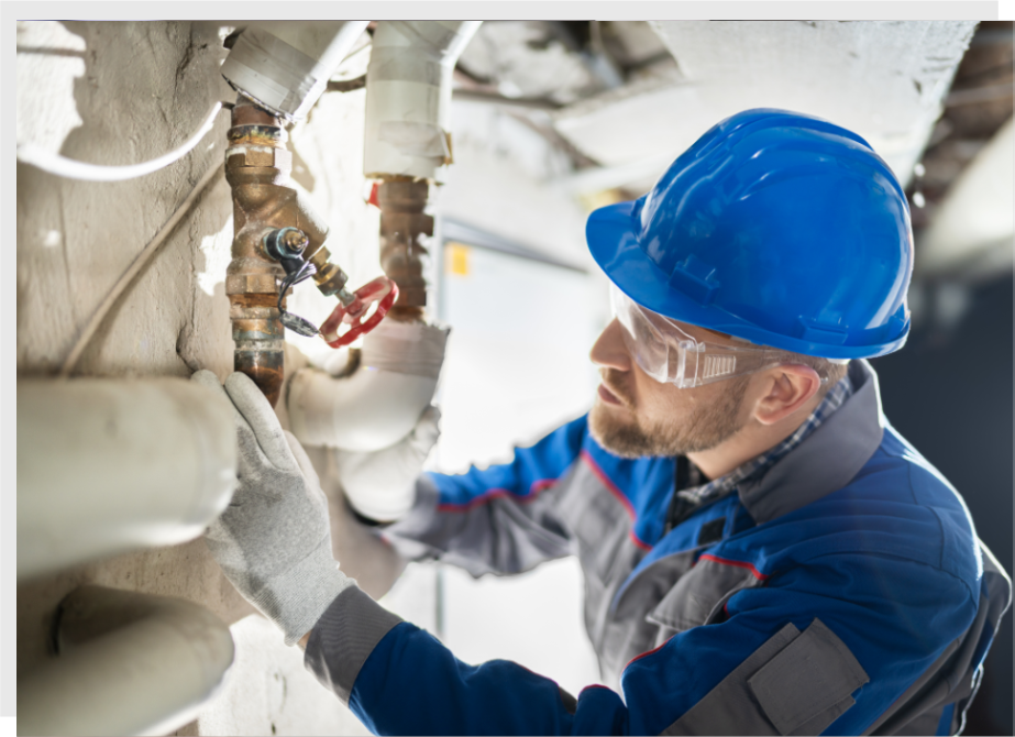 A worker in a blue hard hat and protective goggles inspects pipes in an industrial setting.