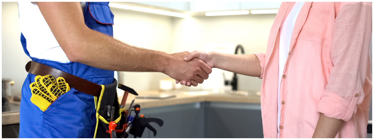 A person in a tool belt and overalls shakes hands with someone wearing a pink shirt in a modern kitchen setting.